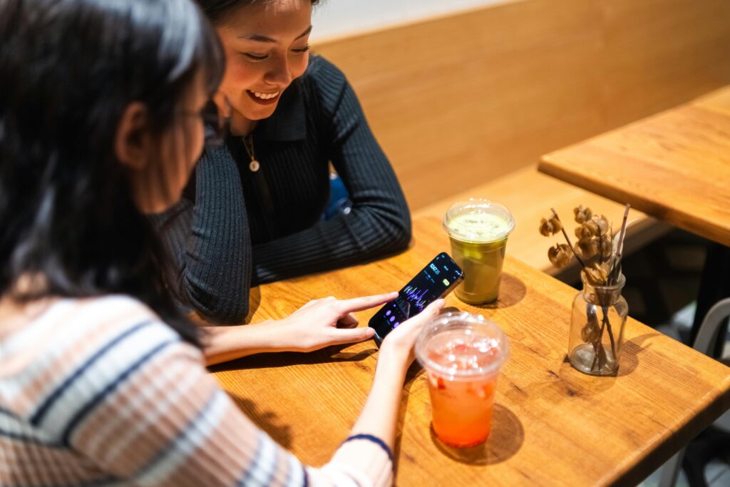 Two friends enjoy drinks at a cafe while looking at a stock market app on a smartphone.